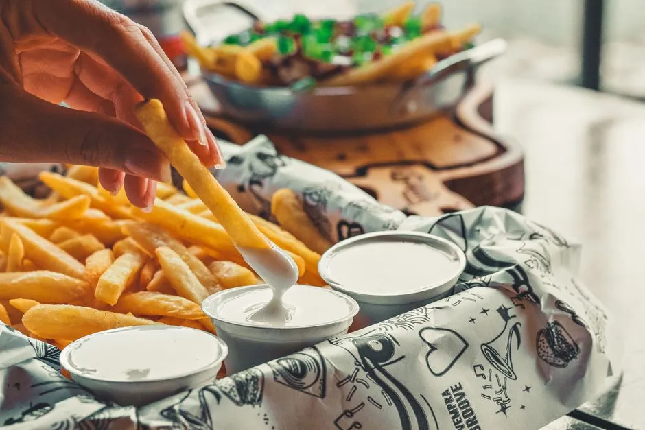 Hand dipping french fry into creamy sauce surrounded by fast food snacks.