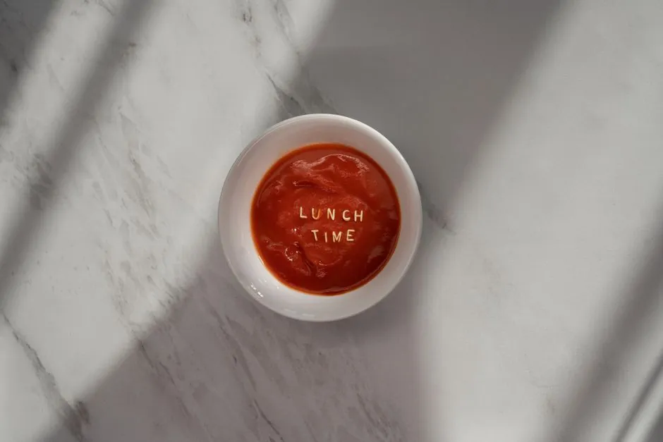 Simple flat lay photograph featuring a bowl of tomato soup with 'lunch time' text on a marble surface.