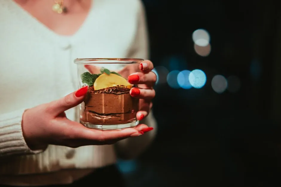 Elegant glass of chocolate mousse dessert in a cozy setting, held by a woman with red nails.