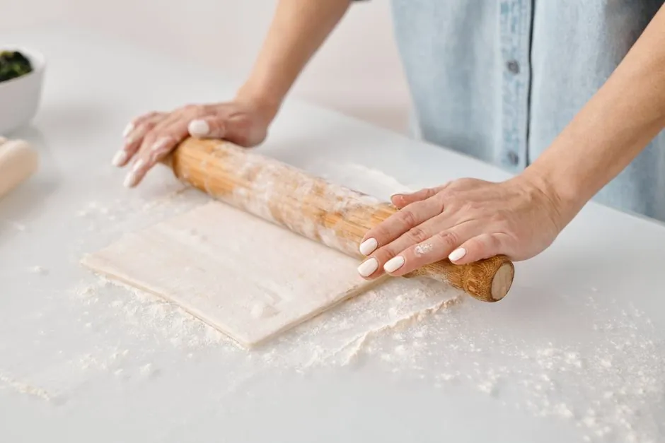 Hands rolling pastry dough on a floured surface with a wooden pin.