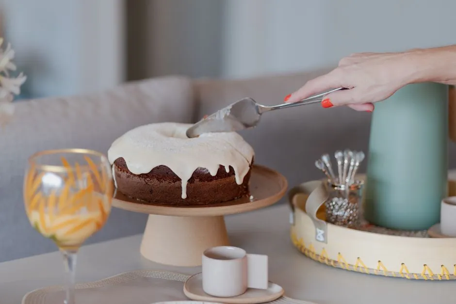 A hand decorates a cake with icing on a beautifully arranged table with drinks and utensils.