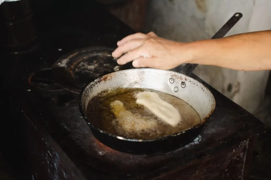 A hand fries traditional bread in oil on a rustic stove, showcasing local culinary techniques.