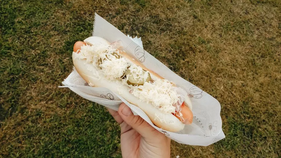 Close-up of a hot dog with cheese and pickles held outdoors on grass.