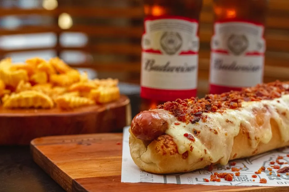 Tasty hot dog topped with cheese and bacon, accompanied by beer bottles and fries on a wooden board.