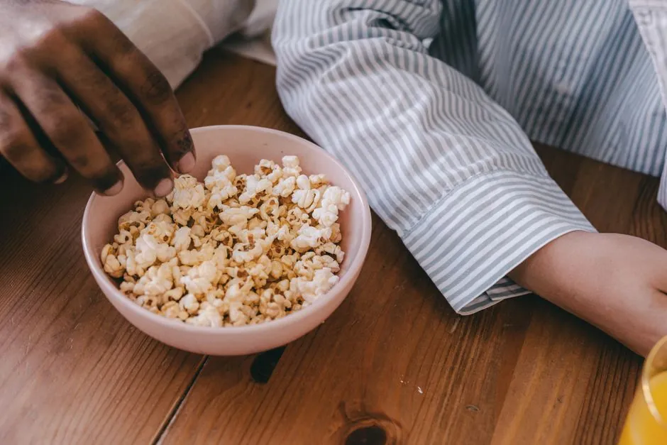 Close-up of hands grabbing popcorn from a ceramic bowl on a wooden table.