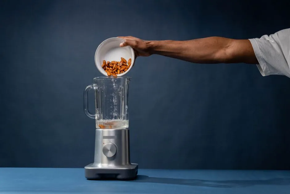 A person adds almonds to a blender, preparing ingredients for a healthy drink in a minimalist studio setting.