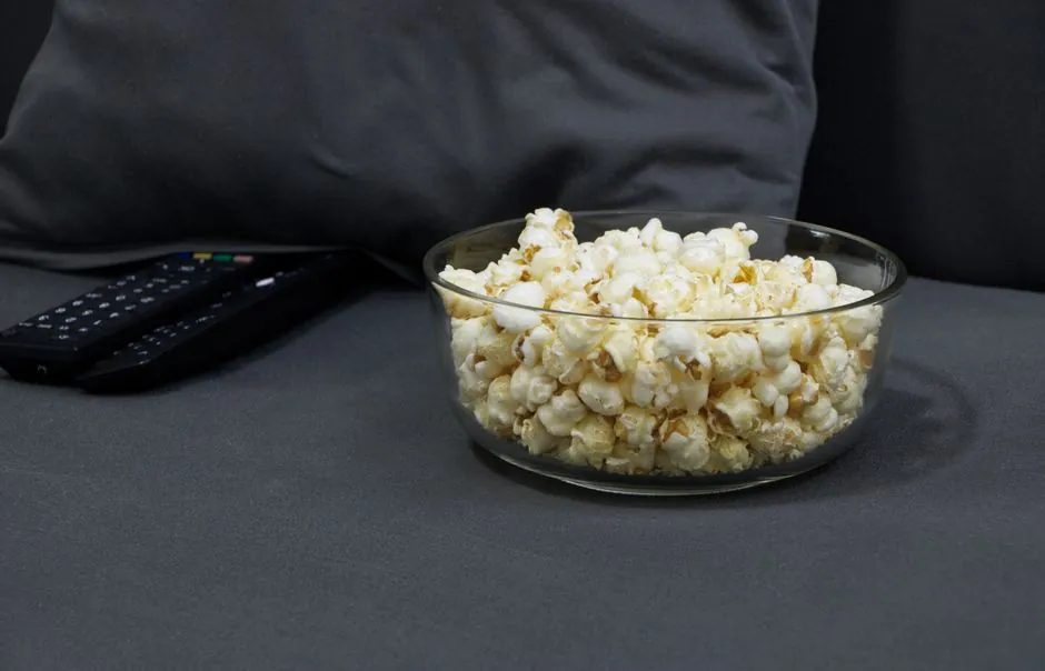 Glass bowl filled with popcorn next to TV remotes on a gray sofa. Perfect for cozy movie nights.